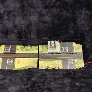 May include: Two sections of model railway track with green and brown scenery, a small building, and a red and black railway crossing sign. The track is connected by a red wire.