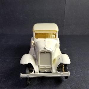 Może przedstawiać: Biały plastikowy model samochodu Ford Model A z lat 20. XX wieku, z czarnym grillem i czarnymi kołami.