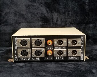 Puede incluir: Una caja beige con un panel negro que presenta múltiples conectores de audio y etiquetas en francés. Las etiquetas incluyen "Droit Gauche", "Radio", "Aire", "12V", "Ampli" y "All".