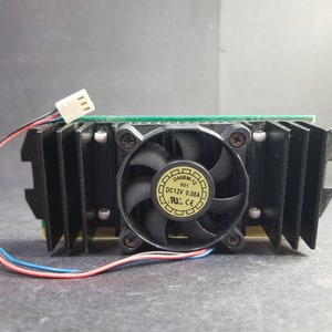 May include: A computer graphics card with a black heatsink, a small fan, and a green circuit board. The fan has a yellow sticker with black text. Red and blue wires extend from the card.