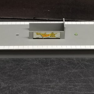 May include: A grey plastic model train station platform with a white trim and a sign that reads "Triang Ships Mini-Mo" in yellow and red letters.
