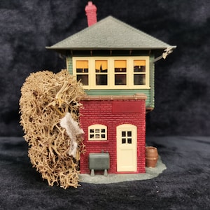 May include: A miniature model of a red brick building with a green roof and a small brown barrel. The building has a small door and windows. The model is surrounded by a brown, dried, moss-like material.