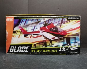 May include: A red, white and black Blade MCX2 helicopter model in a box. The box has the words "Blade #1 by Design MCX2" on it. The box is orange and black.
