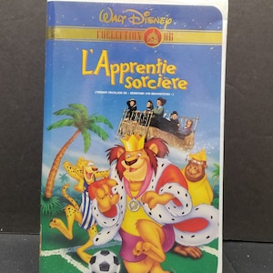 Op de afbeelding: Een VHS-band van de Walt Disney Collection met de film "L'Apprentie Sorcière" in een Franse versie door Rednors en Broomsticks. De hoes toont een cartoon-leeuw die een kroon en een voetbaltenue draagt en voetbal speelt met een cheetah en een aap.