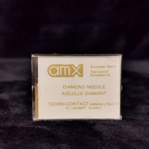 May include: A close-up of a white box with gold lettering. The box contains a diamond needle for a record player. The text on the box reads "amx European Made Fabrication Europeenne DIAMOND NEEDLE AIGUILLE DIAMANT TECHNI-CONTACT CANADA LTEE LTD ST. LAURENT, QUEBEC"