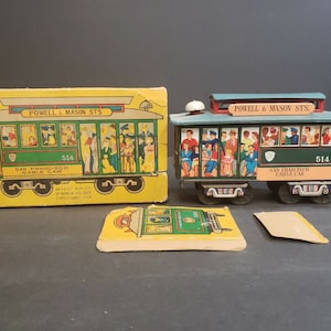 May include: A vintage toy replica of a San Francisco cable car, painted green and yellow with red accents. The toy is in its original box, which features a picture of the cable car and the words "Powell & Mason Sts." The box also includes a small cardboard cutout of the cable car.