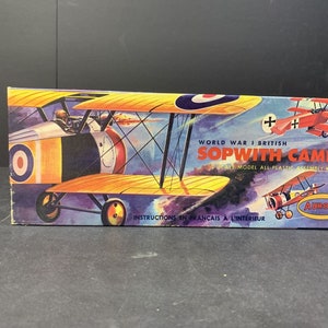 以下が含まれることがあります： 第一次世界大戦のイギリスの航空機、ソプウィス・キャメルをフィーチャーしたビンテージの模型飛行機キットの箱。箱のイラストは、側面に赤、白、青の丸い紋章が付いた飛行機が飛行している様子を描いています。箱には「World War I British Sopwith Camel Scale Model All-Plastic Assembly Kit」と「Aurora」のラベルが貼られています。