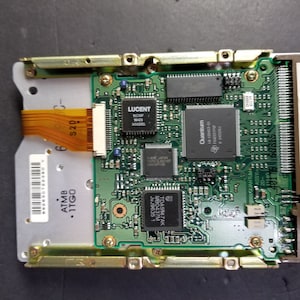 May include: Close-up of a green circuit board with several chips, including a Lucent and a Quantum chip. The board has a gold-coloured connector on the right side and a ribbon cable connected to the left side. The text "1280AT P/N TM124012 Bev 01-B" is printed on the board.