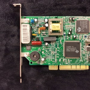 Puede incluir: Una placa de circuito verde con varios componentes electrónicos, incluyendo chips y condensadores. La placa tiene un soporte metálico e incluye texto como "11229-12" y "HONG KONG".