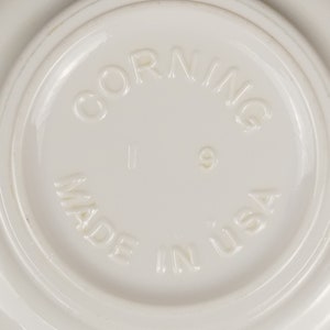 Pode incluir: Close-up da parte inferior de um prato branco Corningware. As palavras "Corning" e "Made in USA" estão gravadas em relevo na superfície branca.