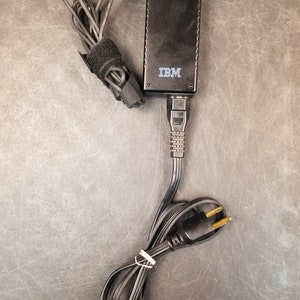 May include: Black laptop power adapter with a black cord and a two-prong plug. The adapter has the word "IBM" printed on it.