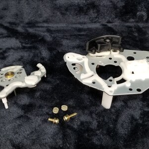 May include: A disassembled silver and white metal part with a black plastic piece. There are four small gold screws on a black surface.