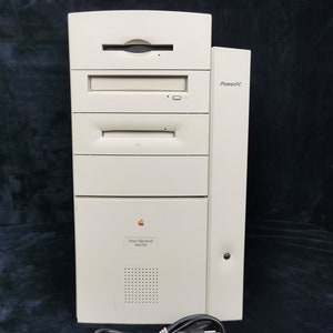 Op de afbeelding: Een beige Power Macintosh 8600 tower-computer met een ingebouwde floppy drive, een CD-ROM-drive en een stroomkabel. De computer heeft een zwart Apple-logo en de tekst "Power Macintosh 8600" aan de voorkant.