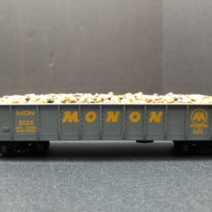 May include: A gray model train car filled with small, light-colored rocks. The car has the word "MONON" in yellow lettering on the side, along with the Monon Railroad logo. The car is on a black surface.