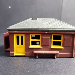 Op de afbeelding: Een klein, bruin plastic model van een gebouw met een grijs dak, een gele deur en ramen, en een bank ervoor. Het model is waarschijnlijk voor een treinset.