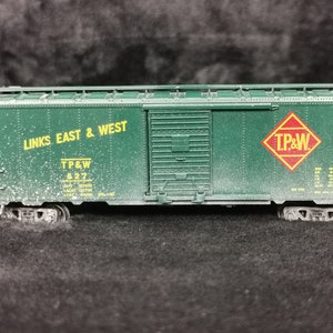 May include: Green model train boxcar with yellow lettering that reads "Links East & West" and "TP&W 627". A red diamond with yellow lettering that reads "TP&W" is on the side of the boxcar.