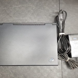 Peut inclure: Un ordinateur portable noir avec un logo IBM argenté en bas à droite. L'ordinateur portable est fermé et repose sur une surface grise. Un adaptateur secteur noir et un cordon sont également visibles sur l'image.