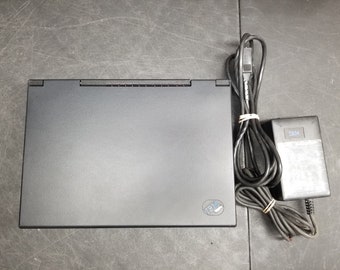 Puede incluir: Un ordenador portátil negro con un logotipo IBM plateado en la esquina inferior derecha. El portátil está cerrado y se encuentra sobre una superficie gris. También se puede ver un adaptador de corriente negro y un cable en la imagen.