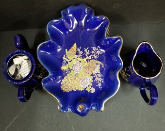 May include: A cobalt blue porcelain serving set with a leaf-shaped dish and two creamer pitchers. The dish features a gold-painted peacock design with floral accents.