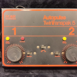 May include: A black and red Troller Autopulse TwinTranspak 5 model train controller with two knobs, each with a red overload light and a yellow track voltage light. The controller has a power switch and a direction switch for each track.