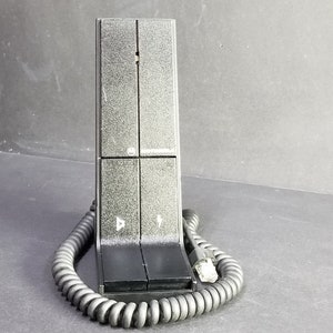 Könnte beinhalten: Ein schwarzes Telefonhörer mit einem Spiralkabel und einer strukturierten Oberfläche. Der Hörer hat zwei Tasten mit Pfeilen darauf.