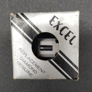 Może przedstawiać: Srebrne i czarne pudełko z nadrukiem "EXCEL" w kolorze czarnym. Pudełko zawiera zamienny diamentowy stylet do gramofonu. Na pudełku widnieje napis "REPLACEMENT DIAMOND GENUINE".
