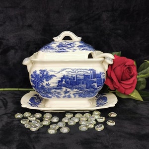 May include: A white and blue porcelain soup tureen with a lid and a matching serving plate. The tureen features a blue and white landscape design with a castle and trees. The tureen is surrounded by clear glass beads.