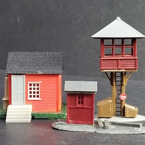 May include: Three miniature red and grey buildings with black roofs. The building on the left has a grey door and a window. The building in the centre has a window. The building on the right is a tower with a sign that reads "Chaffan's Lane".