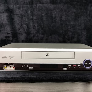 Peut inclure: Un magnétoscope VHS argenté avec un panneau frontal noir. Le lecteur a un logo Z sur le devant et le texte "VHS" en bas à droite.