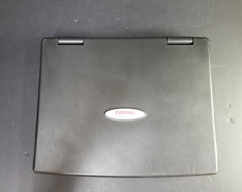 Puede incluir: Un ordenador portátil negro con un logotipo Compaq plateado en la tapa.