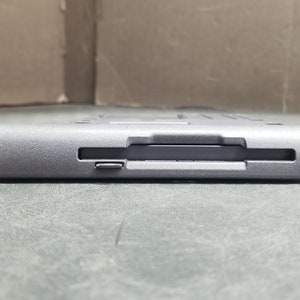 Op de afbeelding: Een grijs, rechthoekig, plastic apparaat met een sleuf voor een geheugenkaart. Het apparaat heeft een getextureerd oppervlak en een kleine knop aan de zijkant.