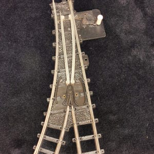 May include: A model train track switch, featuring two diverging tracks. The track is made of silver-colored metal rails and dark gray ties. A black control box with a white lever is attached. This item is designed for model train enthusiasts.