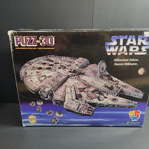 Puede incluir: Una caja de modelo Puzz-3D Star Wars Millennium Falcon. La caja presenta una imagen detallada de la nave espacial Millennium Falcon, con el logotipo de Star Wars en la parte superior. La caja también incluye el texto "857 piezas".