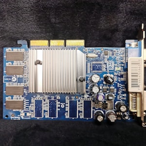 Può includere: Una scheda grafica per computer blu e argento con un dissipatore di calore e un connettore argentato sul lato. La scheda ha un numero di serie 7786561038631V stampato sul lato.