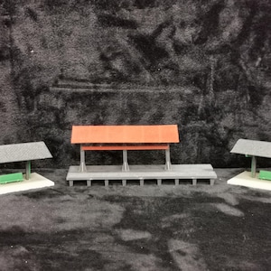 Op de afbeelding: Drie miniatuur treinstations perrons met banken. De perrons zijn gemaakt van grijs plastic en hebben een rood, grijs en zwart dak. De banken zijn groen.