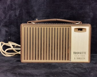 Może przedstawiać: Winylowe radio tranzystorowe w kolorze brązowym i beżowym z uchwytem do przenoszenia. Radio ma wzór imitujący drewno i napis "TRANSETTE SOLID STATE 8 TRANSISTOR" z przodu.