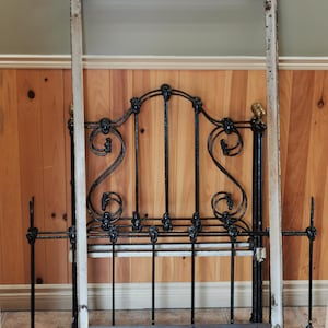 May include: A black metal bed frame with ornate scrollwork and a white painted headboard. The frame is disassembled and lying on a tiled floor.