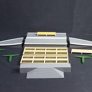 Op de afbeelding: Een modeltreinstationset met een grijs perron, gele banken en groene leuningen. Het station heeft een grijs dak met een reeks ramen en een trap die naar het perron leidt.
