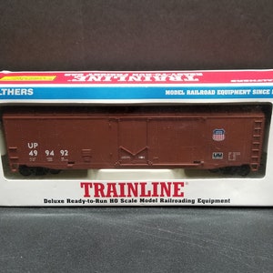 Może przedstawiać: Brązowe pudełko z modelem wagonu kolejowego w środku. Pudełko jest oznaczone "TRAINLINE" i "Deluxe Ready-to-Run HO Scale Model Railroading Equipment". Wagon jest wagonem towarowym z literami "UP" i numerami "49 94 92" na boku.