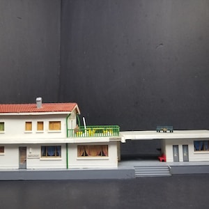 May include: A detailed model of a building with a red tiled roof and white walls. The model features a restaurant sign, windows, and a balcony with green railings. A separate structure with a flat roof and a red bench is attached. The model is on a grey base.