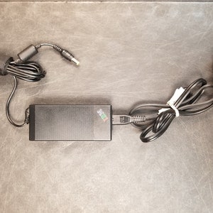 May include: A black laptop power adapter with a black power cord and a black plug. The adapter has a green and white logo on it.