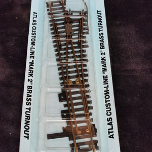 May include: A close-up of a #4 right hand turnout track for model trains. The track is made of brass and is part of the Atlas Custom-Line "Mark 2" series. The packaging includes the text "#4 Right No Brass Turnout #232 Mark 2" and "Atlas Custom-Line "Mark 2" Brass Turnout".