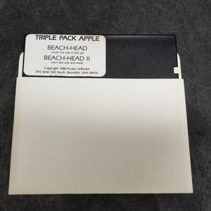 May include: A white floppy disk sleeve with a black label that reads "Triple Pack Apple Beach-Head (insert this side of disk up) Beach-Head II (turn disk over and insert) Copyright 1988 Access Software 545 West 500 South Bountiful, Utah 84010"