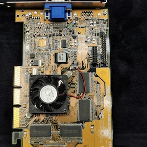 Puede incluir: Primer plano de una placa de circuito impreso de color dorado con un conector VGA azul, un ventilador y el texto "P-V8800" impreso en el lateral. La placa tiene una variedad de chips y otros componentes.