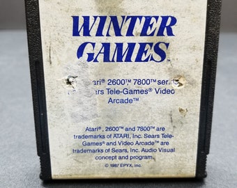 Puede incluir: Un cartucho de juego en blanco y negro para las consolas de videojuegos Atari 2600 y 7800. El cartucho está etiquetado como "EPYX Winter Games" y presenta el texto "Atari, 2600™ y 7800™ son marcas comerciales de ATARI, Inc. Sears Tele-Games® y Video Arcade™ son marcas comerciales de Sears, Inc. Concepto y programa audiovisual. © 1987 EPYX, Inc."