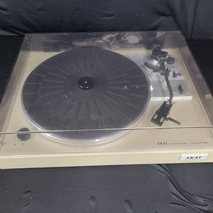 Może przedstawiać: Gramofon Akai AP-D21 w kolorze beżowym z przezroczystą osłoną przeciwkurzową. Gramofon ma czarny talerz i srebrne ramię. Logo Akai jest widoczne na przedniej części gramofonu.