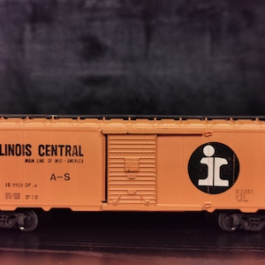 Op de afbeelding: Een oranje modeltreinwagen met de woorden "Illinois Central" en "Main Line of Mid-America" erop gedrukt. De wagen heeft een zwart dak en zwarte wielen. De wagen heeft een zwarte cirkel met de letters "IC" in wit aan de zijkant.