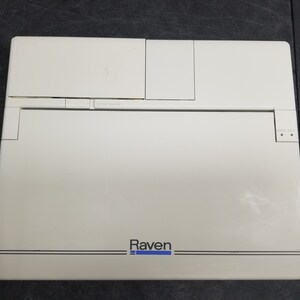 May include: A white Raven laptop computer with a black and blue logo. The laptop has a built-in trackpad and a few buttons on the top.