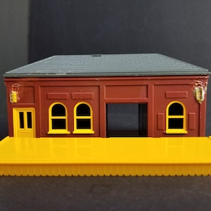 May include: A red and gray plastic model train station with a yellow platform. The station has three windows with yellow frames and a door with a yellow frame. The platform has a scalloped edge.