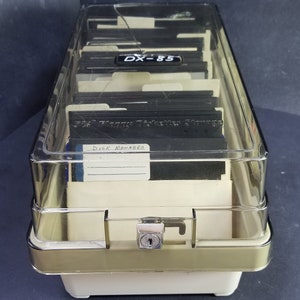 Puede incluir: Una caja de almacenamiento de plástico transparente con un borde dorado y una cerradura. La caja está llena de disquetes. La etiqueta en la parte delantera de la caja dice "DISK MANAGER".